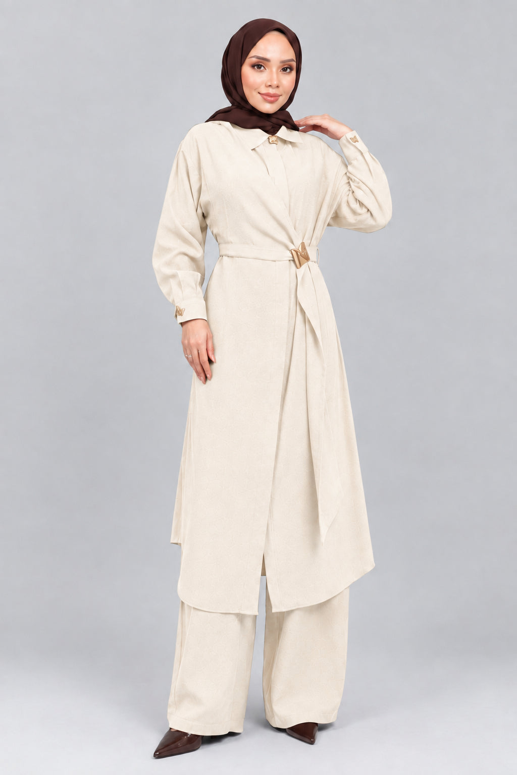 A woman wearing a long brown abaya with a matching headscarf, wide-leg pants, and holding a small brown handbag, standing against a gray concrete wall.