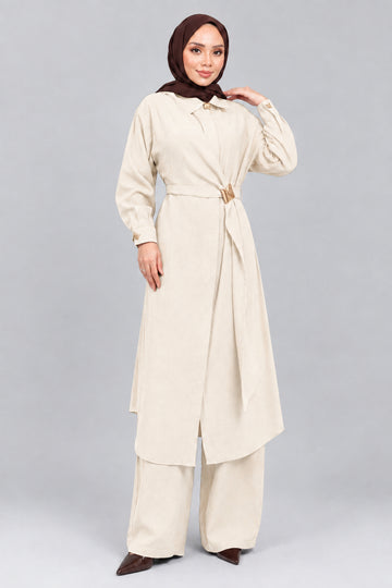 A woman wearing a long brown abaya with a matching headscarf, wide-leg pants, and holding a small brown handbag, standing against a gray concrete wall.