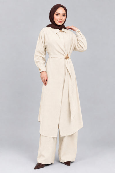 A woman wearing a long brown abaya with a matching headscarf, wide-leg pants, and holding a small brown handbag, standing against a gray concrete wall.