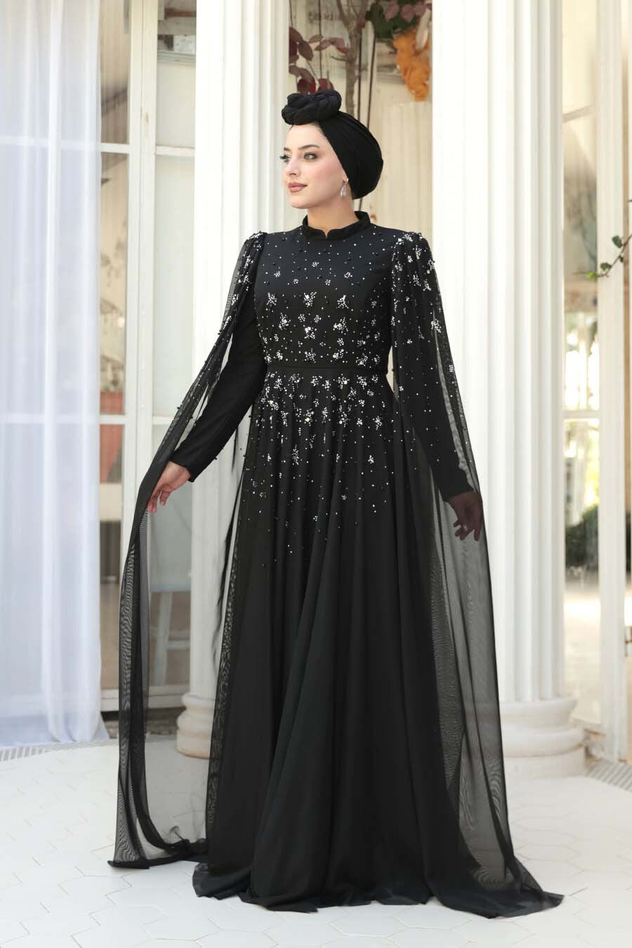 Woman wearing a black evening gown with long sleeves and a matching headscarf indoors.